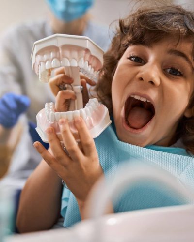 Curly boy opening his mouth wide while holding a human jaws prop while sitting in dentist office